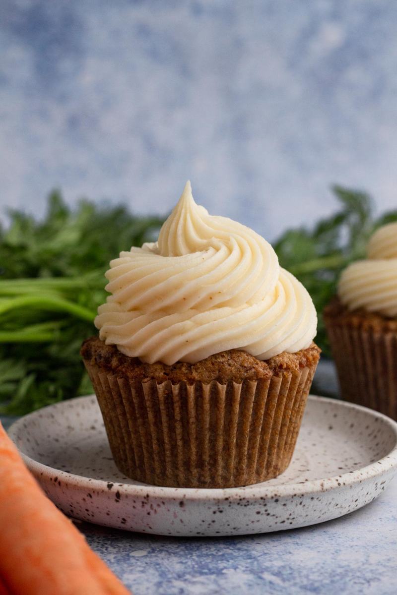 Carrot cake cupcake crowned with tall creamy cream cheese frosting swirl, moist spiced crumb, and nutty garnish