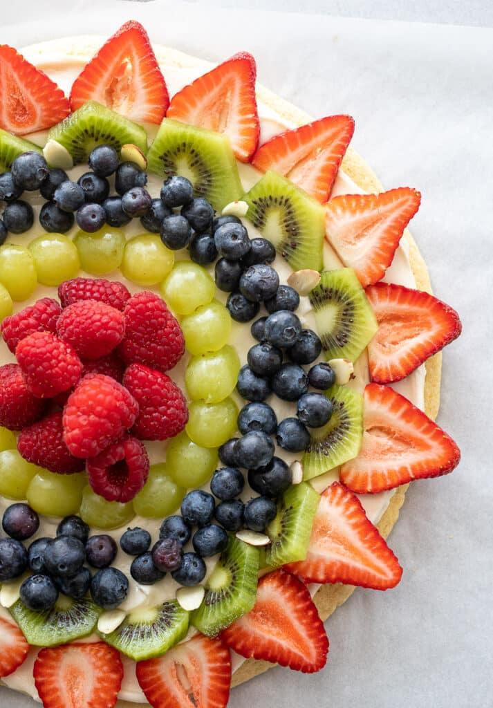 Sugar cookie fruit pizza with cream cheese frosting, white chocolate drizzle, fresh raspberries, kiwi, and blueberries