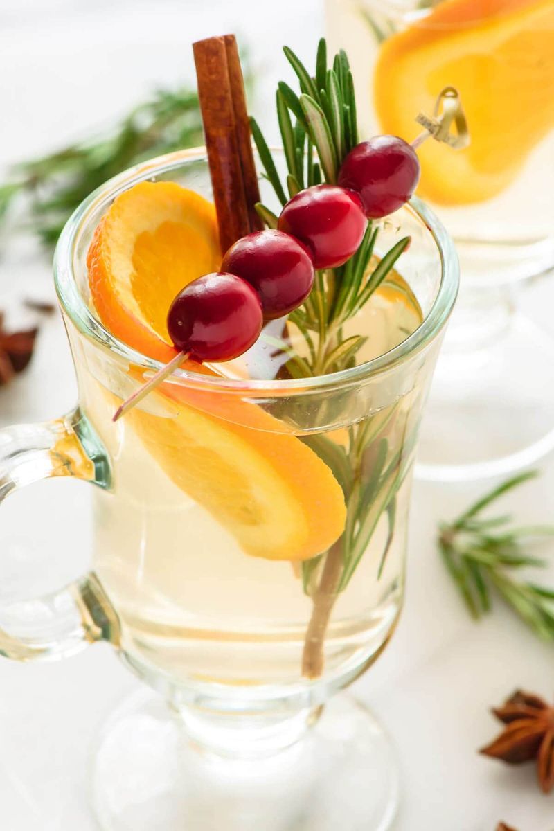 Herbal mulled white wine garnished with fresh thyme sprigs, orange slices, and cinnamon in clear mugs.
