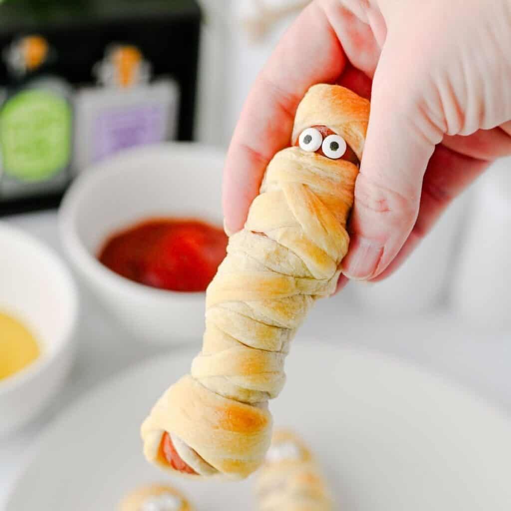 A platter of fully wrapped mummy hot dogs with mustard eyes and a drizzle of ketchup, surrounded by orange “fire” sauce.