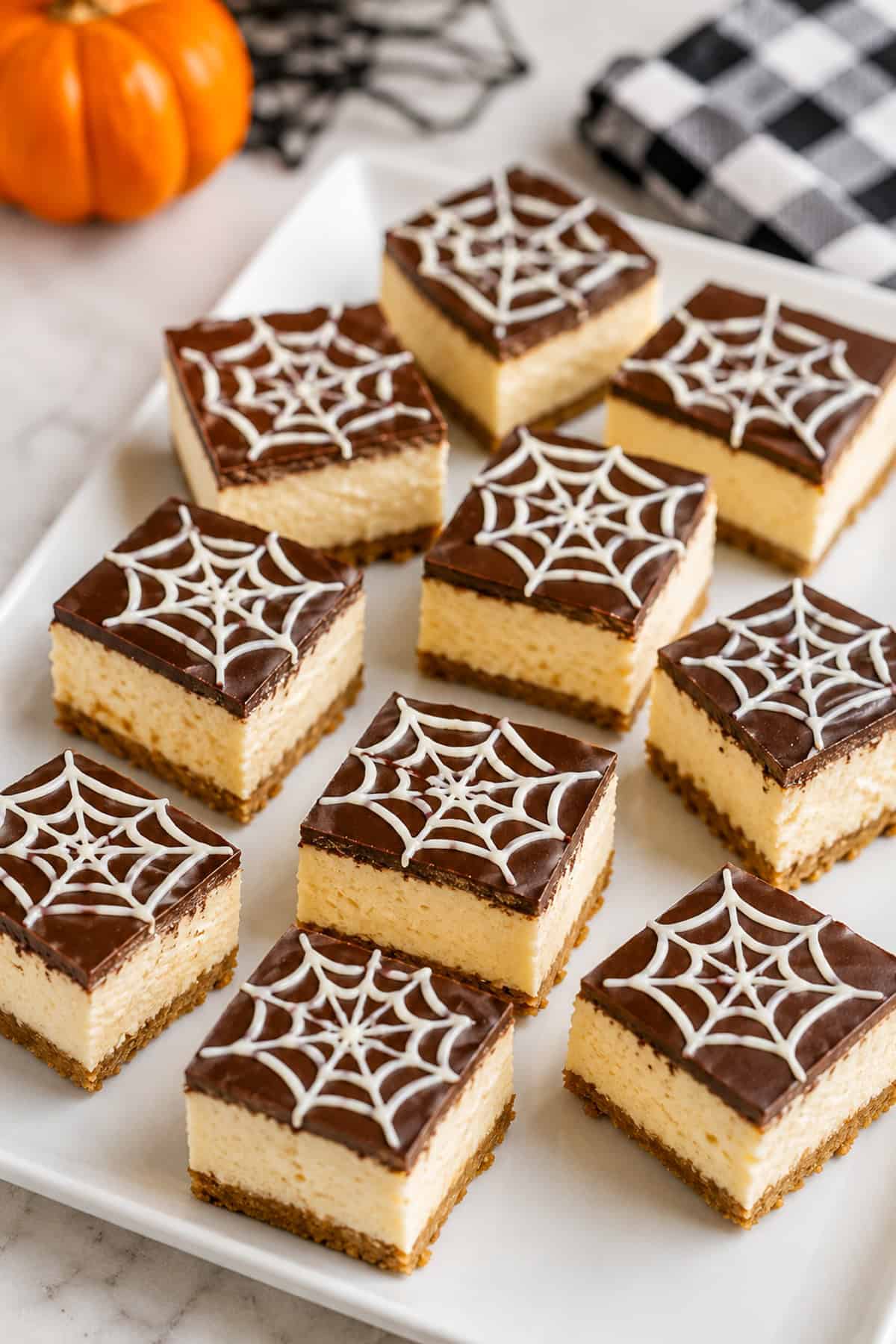 Square chocolate cheesecake bars topped with white chocolate spiderweb designs and red candy eyes on a plate.