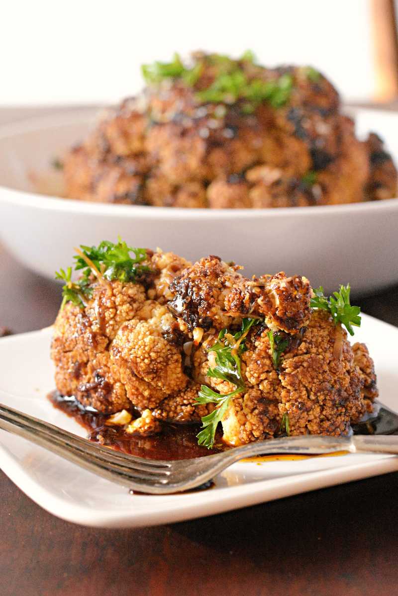 Whole head of roasted cauliflower with charred edges and dark balsamic glaze drizzled over the top with fresh herbs.